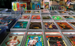 An extensive collection of sports trading cards from the 1990s, displayed on a flat surface. The collection includes numerous PSA graded cards, prominently encased in protective holders. Additionally, there are several rare insert cards and a few unopened boxes of trading cards, suggesting they are mint condition collectibles. The items are arranged to showcase their variety and rarity