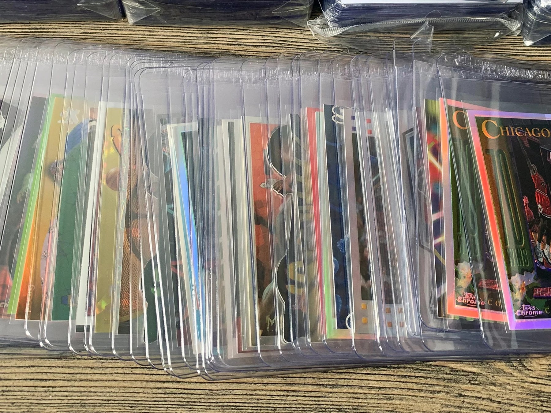 A collection of approximately 100 trading cards from the 1990s, including both insert and refractor cards, displayed in a fanned-out arrangement on a table. Each card is securely housed in either a Card Saver 1 holder or a Card Capsule semi-rigid holder