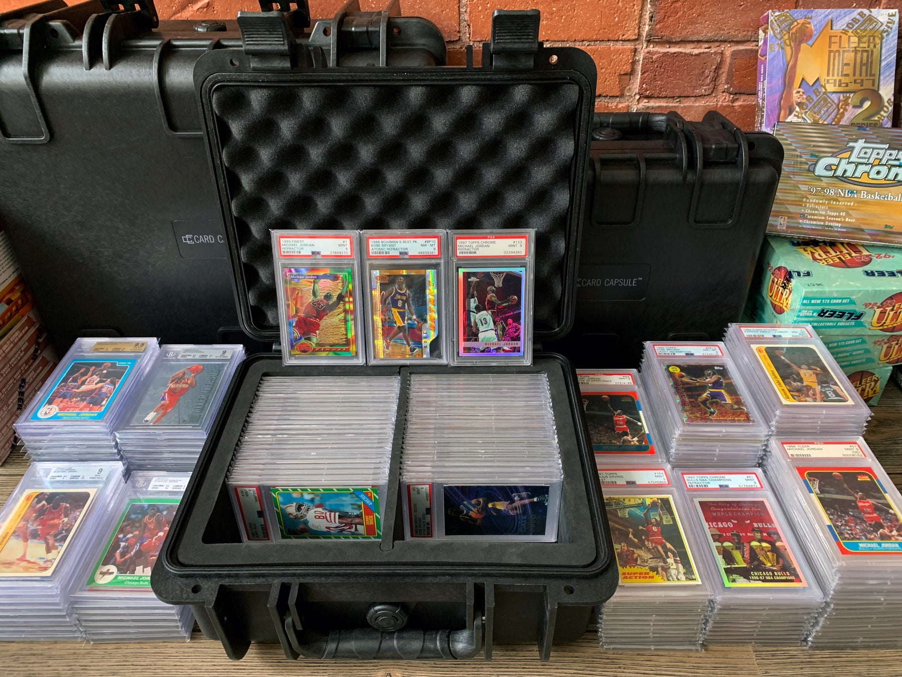 A Card Capsule Genesis waterproof case designed for PSA graded cards is placed on a table surrounded by piles of PSA slabbed cards and various other Card Capsule products
